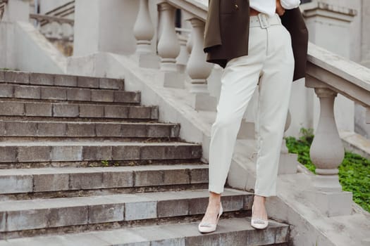 Female fashion outfit. Woman in white trousers and high-heeled shoes posing on stairs outdoors. Crop view on legs