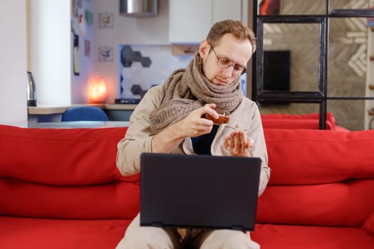 Man in scarf with cold takes medication while sitting on couch with laptop online communication with the attending physician