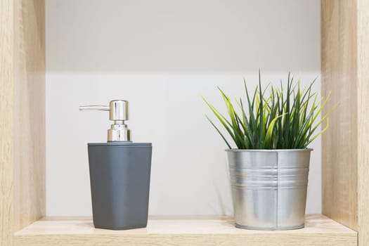 Soap dispensers and artificial houseplant in metal decorative pot on wooden shelf in bathroom. Interior details