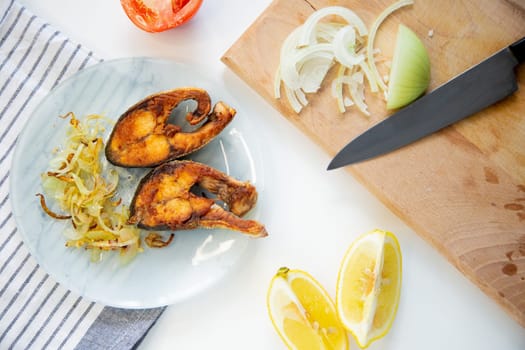 Fried fish with onions is on a plate, on a waffle towel, next to it is a cutting board with chopped onions, lemon and tomato on a white background. High quality photo