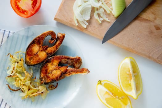 Fried fish with onions is on a plate, on a waffle towel, next to it is a cutting board with chopped onions, lemon and tomato on a white background. High quality photo