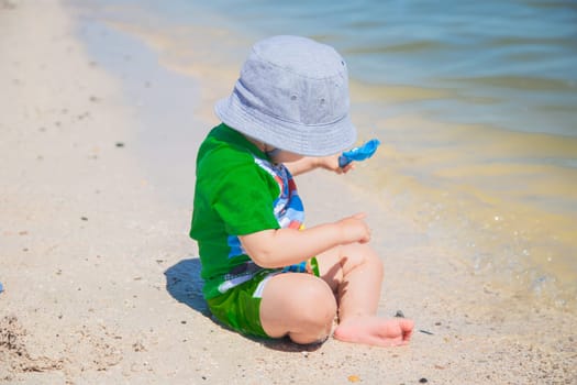 A little boy is sitting in a panama hat on the sand at the water's edge and playing with a shovel. . High quality photo