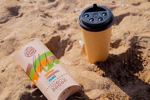 Coffee in a disposable cup and burger from Burger King lie on the sand under the sun. High quality photo