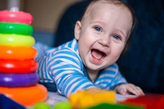 A little kid in a striped suit is lying on his tummy, next to him a children's pyramid and toys. High quality photo