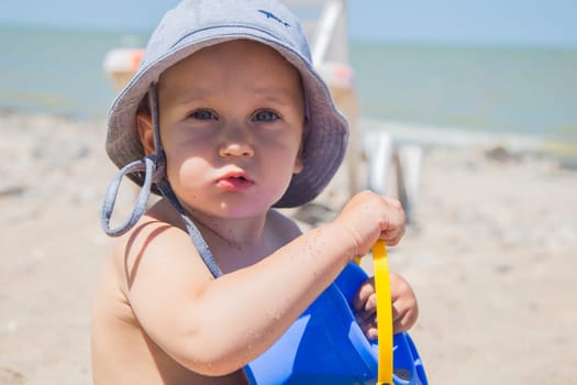 A little boy is sitting in a panama hat on the sea and playing with a bucket close-up. High quality photo