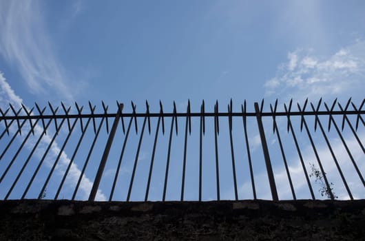 Wrought iron spiky fence placed on the top of a wall. Blue cloudy sky