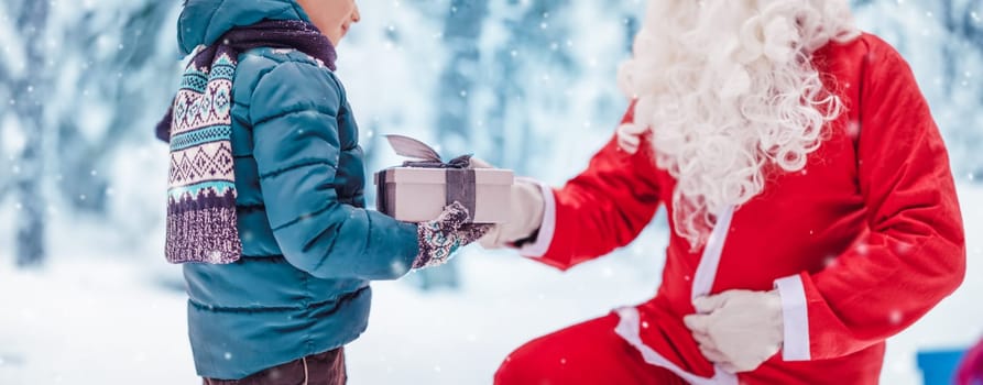 Santa Claus giving present box for child in the snowy park. Concept of the holidays and celebration.
