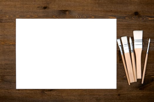 A clean white sheet and brushes on a wood textured background with space to copy. Layout, mockup space for text and drawing.