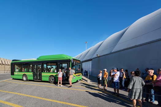 Civitavecchia. Italy - December 19, 2024: Passengers waiting in line next to a green bus at a stop on a sunny day, following the yellow line on the ground. Concept: Organized public transport.