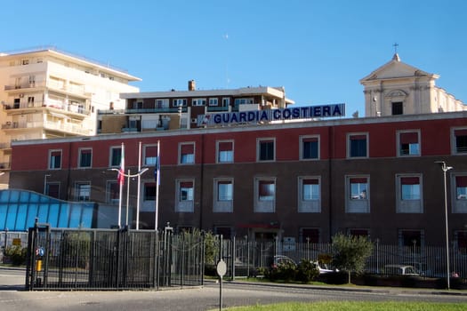 Civitavecchia, Italy - December 19, 2024: Coast Guard building with windows, flags and fenced area stands in front of a church and residential buildings, reflecting maritime security.