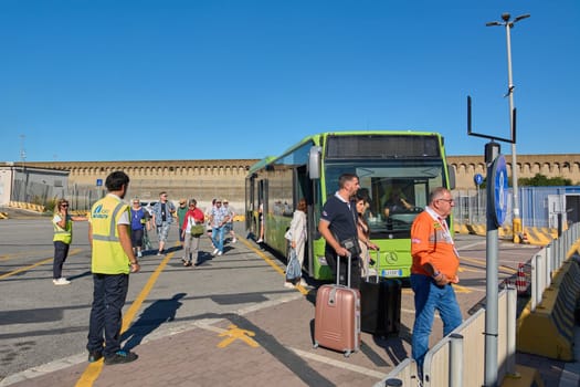 Civitavecchia, Italy - December 19, 2024: Passengers disembark from a bus at a terminal, with staff in high-visibility vests assisting them with luggage and showing travel and transportation services.