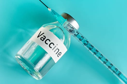 Glass vial with label vaccine and hypodermic syringe needle near, view from above on green cyan background.