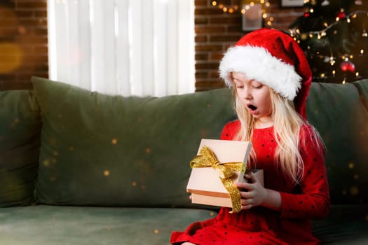 Small little shocked girl in red dress holds Christmas or New Year gift box present. Caucasian child surprised and say wow emotionally, exited because of delivered gifted surprise, expressive emotion