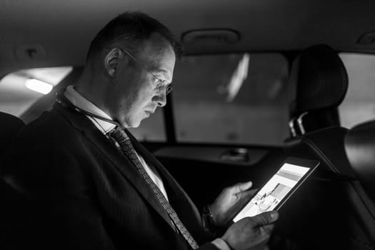 Businessman working remotely on business trip, using digital tablet, sitting in the back seat of taxi car. Black and white image.