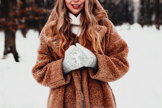 Beautiful young woman in snowy fancy winter woodland. Girl wearing fluffy gloves, cap and coat. Christmas forest, trees on blurred background. Crossed hands because of cold weather. High quality photo