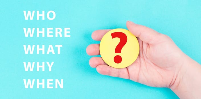 The words who, what, why, when, where are standing on a blue colored background, hand is holding a question mark, brainstorming, searching for answers