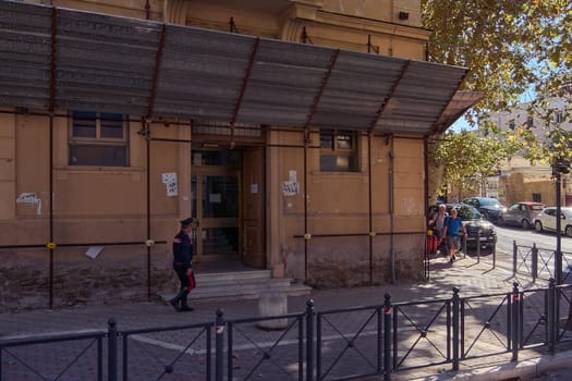 Civitavecchia, Italy - December 19, 2024: Old building with weathered facade and metal awning, with uniformed person walking and cars parked along the street. Concept: Urban history and everyday life.