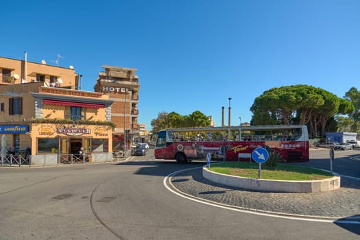 Civitavecchia, Italy - December 19, 2024: An open-top tour bus on a roundabout in Civitavecchia passing buildings with signs indicating a pizzeria and a hotel Concept: urban tourism and transportation