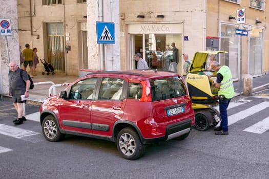 Civitavecchia, Italy - December 19, 2024: A postal worker in a high-visibility vest stands by a yellow Poste Italiane electric vehicle, handling mail. A red Fiat Panda is parked at a crosswalk.