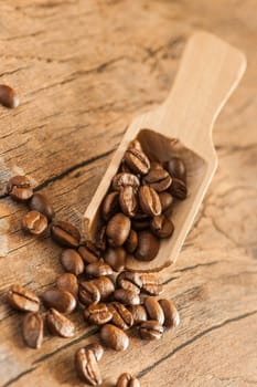 Close-up of roasted coffee beans with a small wooden scoop on a surface made of old wood in a vertical orientation.