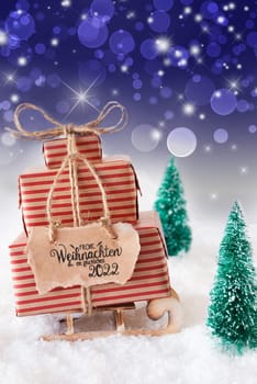 Sled With Christmas Gift And Label With German Frohe Weihnachten Und Ein Glueckliches 2022 Means Merry Christmas And A Happy 2022. Snow With Christmas Tree And Purple Sparkling Background