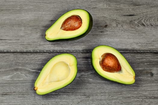 Tabletop view - three halves of cut avocado, some with seed pit, on gray wood desk.