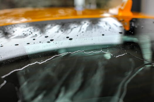 Shallow depth of field (only small drops in focus) yellow car rear window covered with water in carwash / rain.