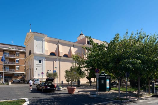 Civitavecchia. Italia  - December 27, 2024: An urban square in Civitavecchia, Italy, with a historical church, trees, and parked cars under a bright sunny sky. Concept: Cityscape and cultural heritage