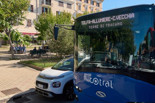 Civitavecchia, Italia - December 27, 2024: Blue public bus and parked car in a sunny square, locals on benches, trees, and vibrant urban scene. Concept: Urban life, transportation, and community.