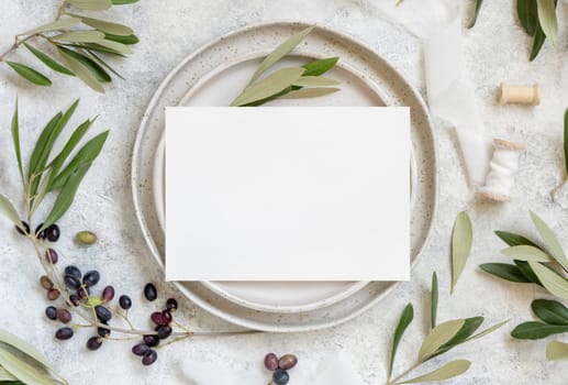 Wedding Table place with invitation card and porcelain plates decorated with olive branches top view. Elegant modern template with horizontal blank paper card flat lay. Mediterranean mockup, copy space