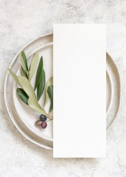 Wedding Table place with a menu card and porcelain plates decorated with olive branches top view. Elegant modern template with vertical blank paper card flat lay. Mediterranean mockup, copy space