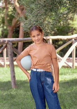 Happy schooler girl in beige t-shirt holding ball outdoors. Cute child doing sports, enjoying playing soccer and having fun in park. Active hobbies, healthy lifestyle concept. T-shirt mockup