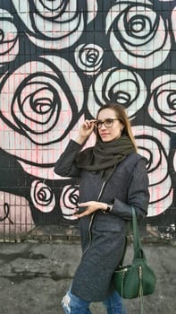 A woman in a grey coat and green scarf with a green bag puts on glasses