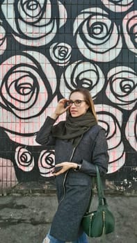 A young woman in a gray coat, green scarf adjusts glasses against the background of a colored wall