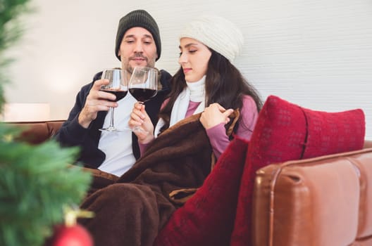 Smiling lovers sitting on sofa and clinking glasses of red wine in living room. Romantic winter holiday and Christmas eve together concept