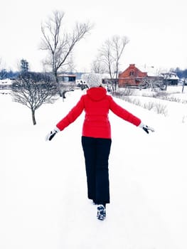 View from the back of a woman in a red jacket, a white hat and black sweatpants walking in the winter through the snow outside the city, winter domestic travel