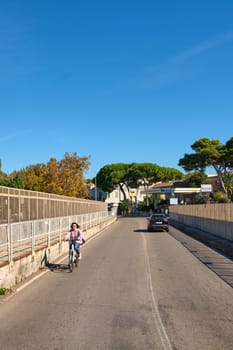 Civitavecchia, Italy - December 27, 2024: Cyclist rides on urban street in Civitavecchia on a sunny day. The road with fences and trees, with a car in the background. Concept: urban transport.