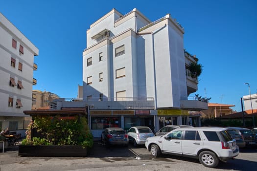 Civitavecchia. Italia - December 27, 2024: Multi-story building with gray facade, windows, and storefronts on ground floor. Parked cars in front and blue sky. Concept: Urban architecture and commerce.