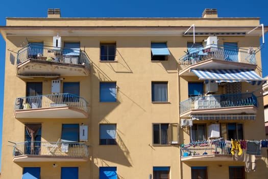 Yellow building with blue shutters and multiple balconies. Balconies have metal railings, awnings, and household items like laundry and chairs. Concept: Residential architecture.