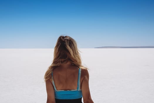 rear view of blonde beautiful Caucasian young woman dressed in blue looking at white salt lake stretching to the horizon with clear sky above