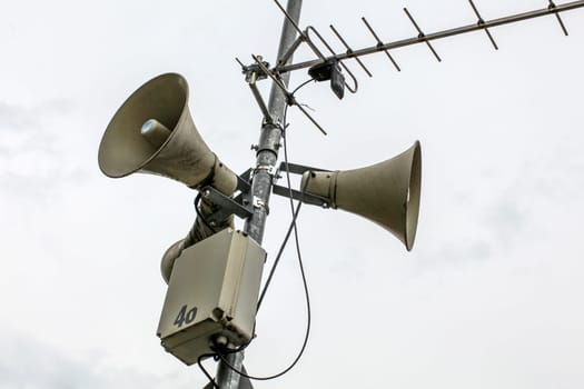 Public address system speakers on a pole, antenna and box with number 46 partially peeled off, overcast sky in background.