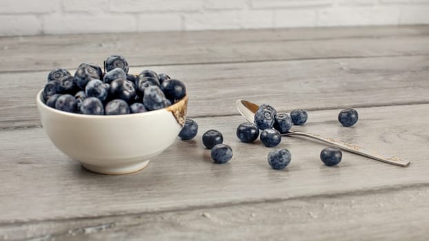 Small bowl of blueberries, some of them spilled next to spoon on gray wood desk.