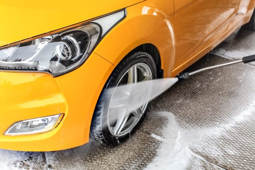Detail on water jet spraying from the hose to yellow car front wheel and tyre, during cleaning in carwash.