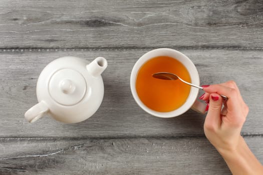Table top view - woman hand holding spoon, stirring tea in white porcelain cup.