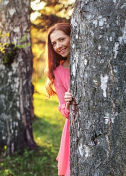 Young woman in pink dress, peek a boo behind the tree, with sunset lit park in background.