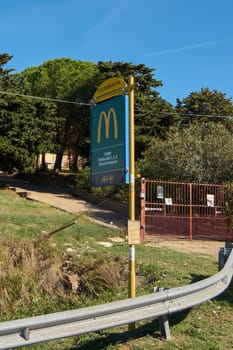 Civitavecchia, Italy - December 27, 2024: Bus stop sign with McDonald's logo and locations, in rural area with vegetation. Metal railing and red door in foreground. Concept: Branding in public spaces.