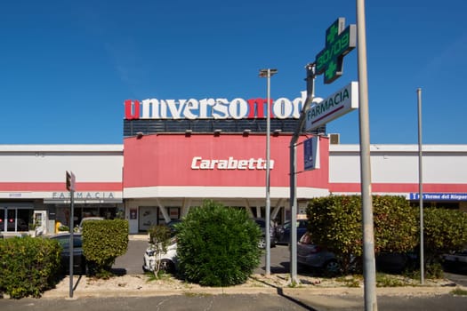Civitavecchia. Italia- December 27, 2024: Exterior of commercial building with universomoda Carabetta,and FARMACIA signs Bushes and parked cars visible under a clear blue sky Concept: Urban commerce.
