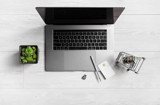 Online shopping concept. Laptop, shopping trolley with dollars, credit card, pen, flash drive and houseplant. Flat lay.