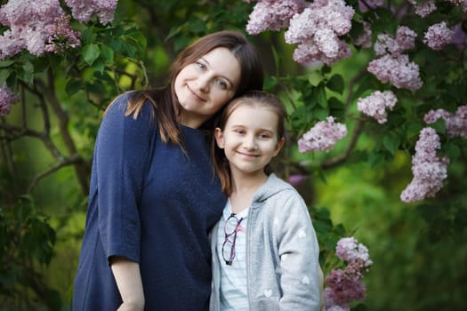 Mom and daughter are posing against the backdrop of a blooming lilac bush.