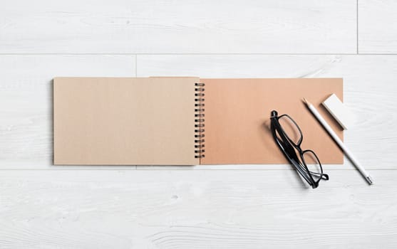 Blank open craft notebook, glasses, pencil and eraser on wooden background. Flat lay.